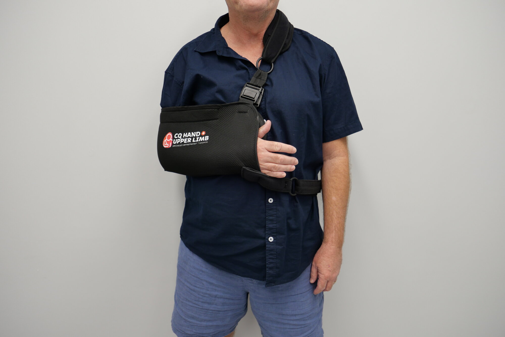 A patient wearing a CQ Hand & Upper Limb branded shoulder sling, forearm supported across the body.